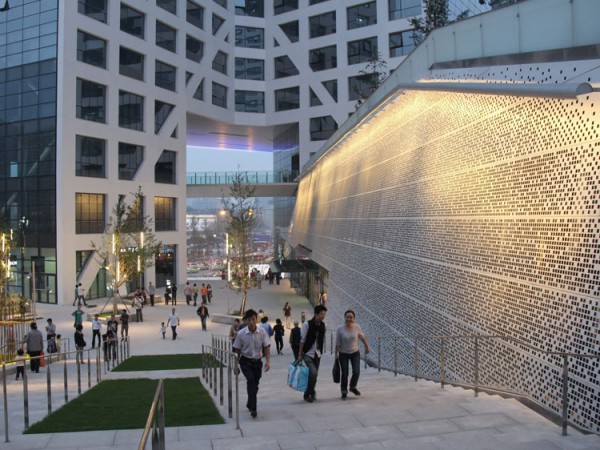 The perforated skin of the central shopping complex is lit up in the evening. Image via Steven Holl Architects.